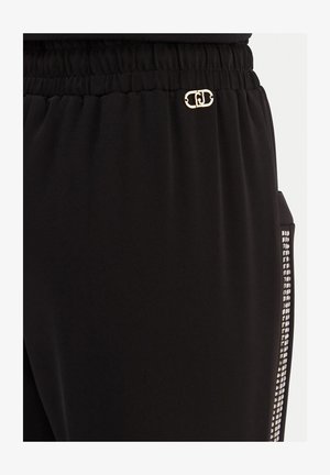 Zwarte broek met elastische tailleband, klein gouden logo bij de taille en een verticale rij zilveren vierkante studs op het zijzakje.