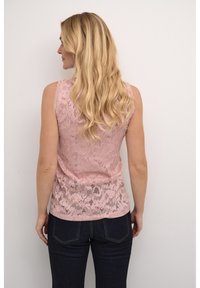 Bez piedurknēm, gaiši rozā mežģīņu tops ar ziedu rakstu, ar brīvu piegriezumu un teksturētu apdari, apvienots ar tumši ziliem džinsiem.