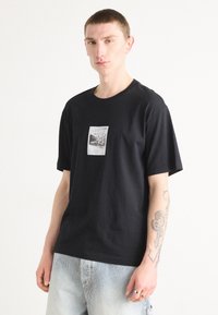 T-shirt nera in cotone con stampa grafica centrale. Design a maniche corte e collo a giro. Il modello indossa anche pantaloni corta in denim a righe.