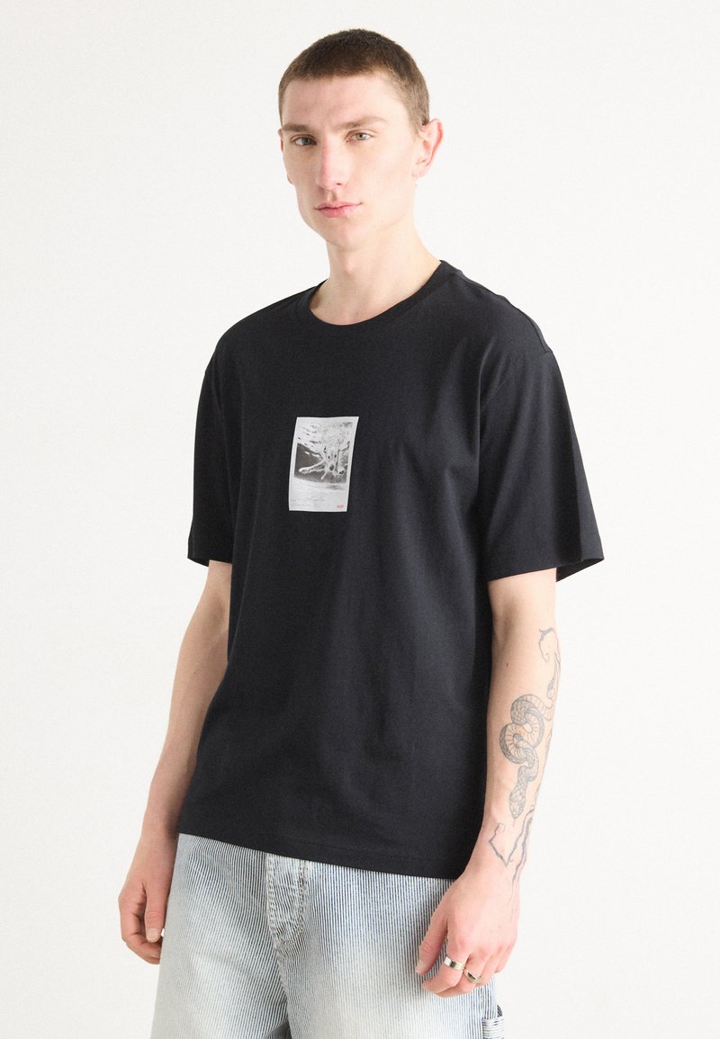 T-shirt nera in cotone con stampa grafica centrale. Design a maniche corte e collo a giro. Il modello indossa anche pantaloni corta in denim a righe.