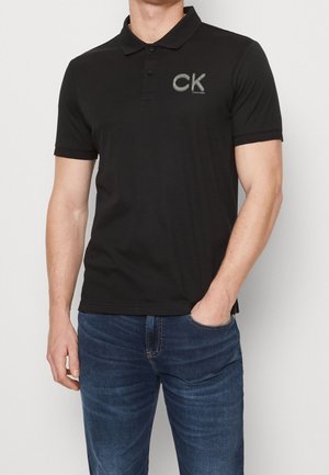 Mies, jolla on musta Calvin Klein -polopaita, jossa CK-logo rinnassa, ja siniset farkut, toinen käsi taskussa yksiväristä taustaa vasten.