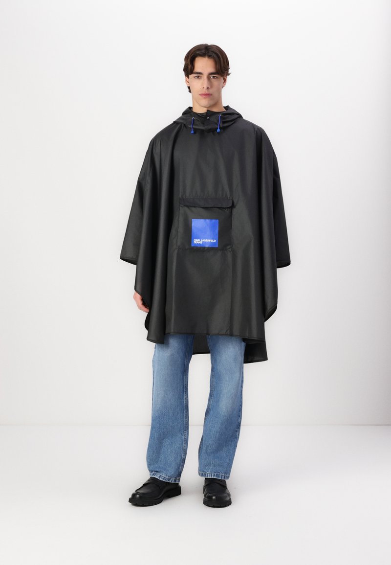 Poncho impermeabil negru cu glugă, având un buzunar frontal și branduire albastră. Textură netedă, croi lejer, combinat cu blugi albaștri deschiși.