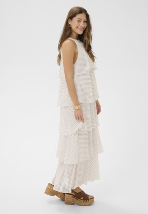 Fiatal nő mosolyog, fehér, réteges ruhát visel, amelyen apró fekete pöttyök vannak, valamint barna platform szandált parafa sarokkal.