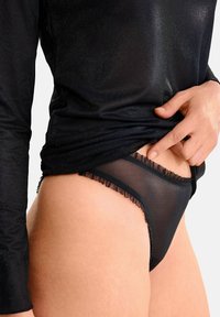 Une personne soulève un haut à manches longues noir scintillant pour révéler un sous-vêtement noir transparent avec des bords froncés au niveau des hanches, contre un fond blanc.