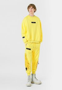 Sudadera amarilla brillante y chándal a juego, con un gráfico de un león en la pierna izquierda, con detalles y etiquetas negras. Completado con botas blancas.