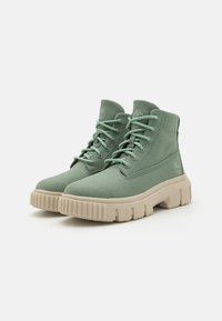 Stivaletti alla caviglia in tela verde con suole spesse beige, caratterizzati da lacci rotondi e dettagli del logo sui lati. Superficie testurizzata e design alto.