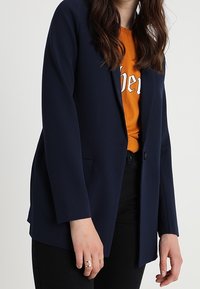 Femme portant un blazer bleu marine sur une chemise orange avec un pantalon noir, se tenant devant un fond clair uni.