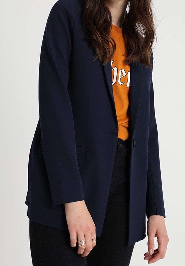 Femme portant un blazer bleu marine sur une chemise orange avec un pantalon noir, se tenant devant un fond clair uni.