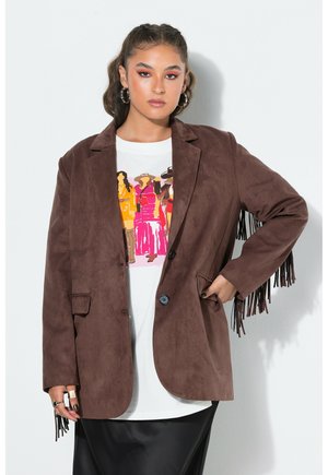 Jeune femme portant une veste en daim marron à franges sur un t-shirt blanc avec un motif graphique et une jupe noire, debout avec une main dans la poche.