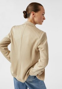 Beiger gestreifter Blazer mit lockerem Schnitt, ausgestattet mit einem Reverskragen, langen Ärmeln und dezenten blauen Nadelstreifen. Getragen über hellblauen Jeans.