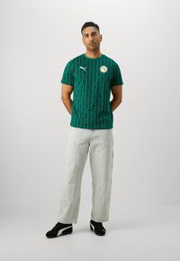 Grünes T-Shirt mit diamantförmigen Mustern, kurzen Ärmeln, weißem Puma-Logo und rundem Abzeichen, kombiniert mit hellblauen, weit geschnittenen Jeans und schwarzen Schuhen.