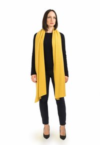 Sciarpa gialla brillante realizzata in tessuto morbido, che si drappeggia elegantemente, abbinata a un maglione scuro e a pantaloni neri aderenti, completata da décolleté a punta.