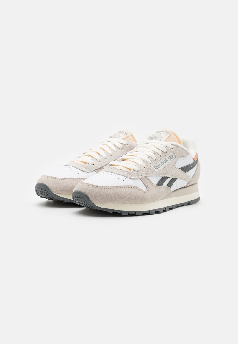 Baskets blanches et beiges en matériaux suédé et en mesh. Présentent des accents gris, des lacets ronds et une semelle en caoutchouc texturé. Marque Reebok visible.