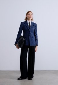 Μπλε σακάκι με ρίγες και δομημένους ώμους, συνδυασμένο με φαρδιά μαύρη παντελόνα και μαύρη τσάντα. Το σύνολο περιλαμβάνει λευκό πουκάμισο και γραβάτα.