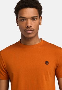 Orange bomulds T-shirt med rund hals, korte ærmer og et lille navy logo på brystet. Glat tekstur og afslappet pasform.