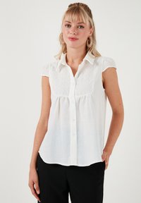 Bílá halenka s krátkým rukávem, která má zapínání na knoflíky, texturovanou krajkovou část, měkký bavlněný materiál a zakřivený lem.
