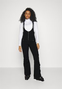 Schwarze Skioveralls mit verstellbaren Trägern, einer vorderen Reißverschlusstasche und einem körperbetonten Design. Kombiniert mit einem weißen Langarmoberteil und schwarzen Stiefeln.