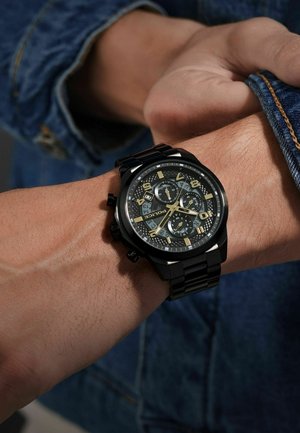 Black chronograph watch with a textured black dial, gold accents, and a matching black metal band, worn on a wrist with visible hair.