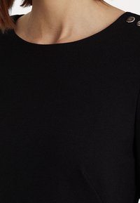 Svart topp med rund halsringning, med metallknappar på axlarna. Tyget ser slätt ut med en mjuk textur.