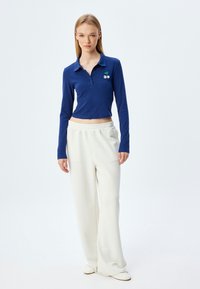 Polo blu a maniche lunghe con colletto e dettaglio dei bottoni, abbinato a pantaloni a gamba larga color crema; presenta un ricamo di ciliegie verdi sul petto.