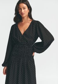 Černé šaty s puntíky, hlubokým výstřihem do V, dlouhými nadýchanými rukávy a nařaseným pasem. Vyznačují se bílými puntíky rovnoměrně rozloženými po celé ploše.