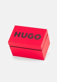 Czerwony prostokątny box z czarnym logo "HUGO" na górze. Gładkie, błyszczące wykończenie i pokrywa na zawiasach. Lekko pochylony dla perspektywy.