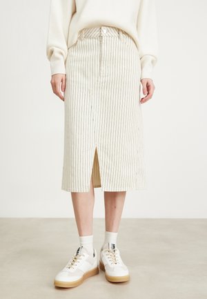 Personne portant un pull crème, une jupe midi crème à fines rayures verticales et fente avant, des chaussettes blanches et des baskets blanches avec des semelles beige.