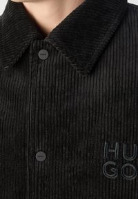 Gros plan d'une veste en velours côtelé noire avec col, deux boutons noirs marqués « HUGO » et logo partiel « HUGO » brodé sur la poitrine.