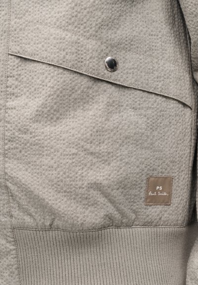 Section de veste en tissu gris clair avec poche à rabat inclinée sécurisée par un bouton argenté et une étiquette PS Paul Smith cousue.