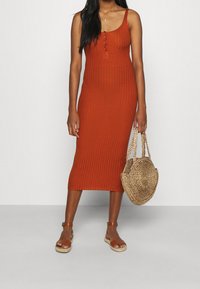Vestito aderente a coste di colore arancio bruciato con scollatura a barchetta e chiusura a tre bottoni. Accompagnato da una borsa rotonda in paglia intrecciata e sandali beige.