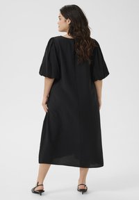 Abito nero in tessuto leggero, con maniche corte a sbuffo e silhouette dritta, dotato di una slit sul retro per facilitare i movimenti.