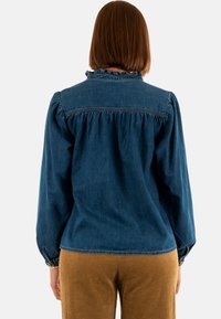 Camicia in denim blu scuro con maniche a sbuffo, colletto arricciato e dettaglio raccolto nella parte posteriore, abbinata a pantaloni in corduroy beige.