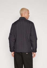 Giacca nera imbottita con motivo a righe verticali, dotata di colletto alto e polsini elastici, abbinata a pantaloni scuri.