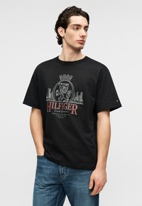 Μαύρο βαμβακερό T-shirt με κοντά μανίκια, διακοσμημένο με γραφική εκτύπωση ενός λιονταριού και αστικού τοπίου, με κόκκινες και λευκές λεπτομέρειες σε κείμενο και λογότυπο.