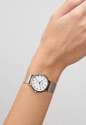 Pols met een horloge met een roségouden meshband, een witte wijzerplaat, zwarte uurmarkeringen en drie wijzers die 10:09:38 aangeven, tegen een lichtgrijze achtergrond.