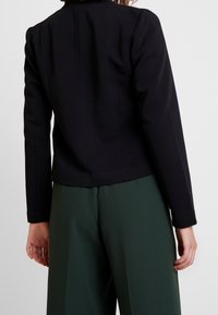 Blazer noir texturé à manches longues, coupe ajustée et longueur raccourcie. Associé à un pantalon vert taille haute, vue de dos.