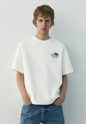 Fiatal férfi rövid, göndör hajjal fehér, túlméretezett pólót visel, rajta a "The Pool Spot" logóval, és kék farmert, egy egyszínű világos háttér előtt áll.