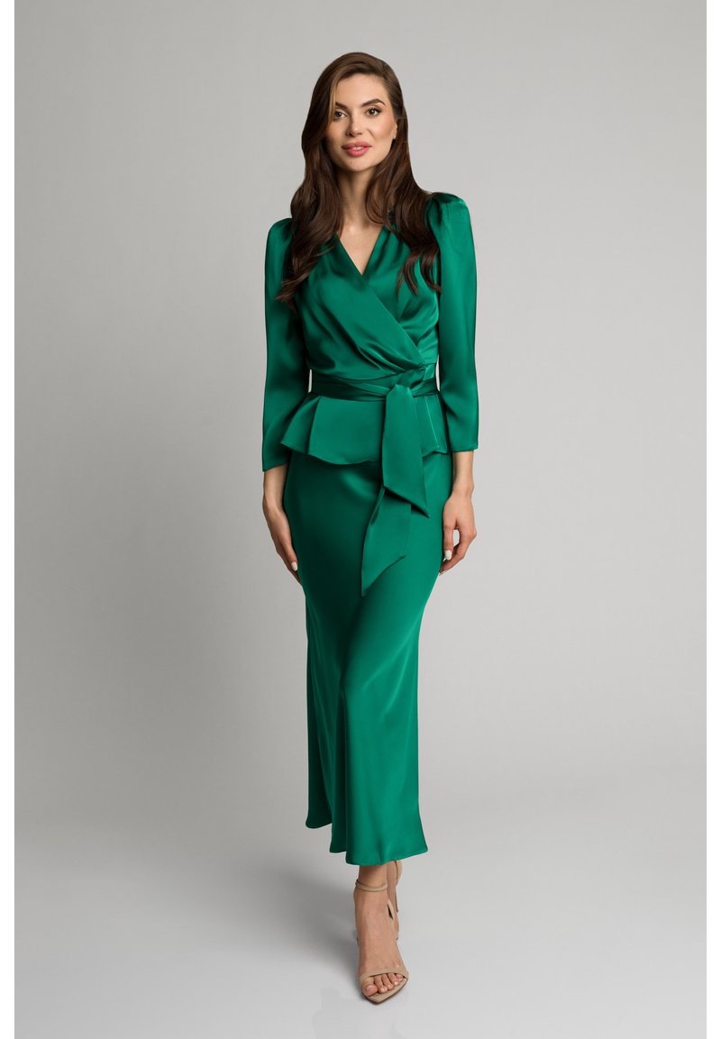 Zielona satynowa sukienka o fasonie kopertowym, z długimi rękawami, w talii peplum oraz zwiewnym kroju. Posiada zawiązany pasek w talii i gładką fakturę.