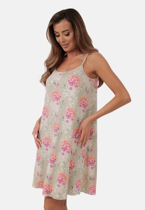 Kobieta o długich brązowych włosach, ubrana w beżową, kwiecistą, bezrękawnikową sukienkę z różowymi i pomarańczowymi kwiatami, stojąca z rękami na biodrach.