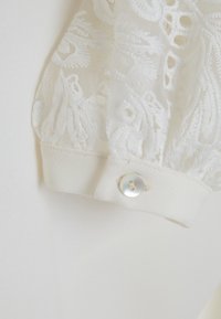 White blouse with sheer embroidered long sleeve, featuring a cuff with a white button. Textured floral pattern in the fabric.