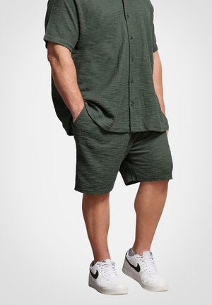 Mand stående med hænderne i lommerne iført en mørkegrøn struktureret kortærmet skjorte med knapper, matchende shorts og hvide Nike-sneakers.