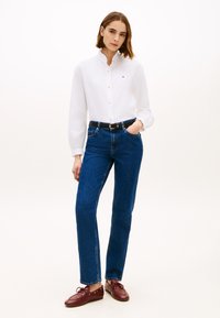 Γυναίκα που φοράει λευκό πουκάμισο με κουμπιά, σκούρο μπλε τζιν με μαύρη ζώνη και μπορντό παπούτσια με κορδόνια, στέκεται με το ένα χέρι στην τσέπη.