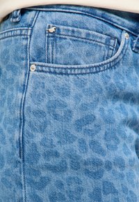 Prim-plan cu blugi din denim albastru deschis, cu un model subtil de imprimeu animal și un mic pin metalic în formă de cățel pe buzunarul din față.