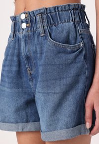 Närbild av blå denimshorts med elastisk midja, dubbel knappstängning, vikta fållar och framfickor som bärs av en person.