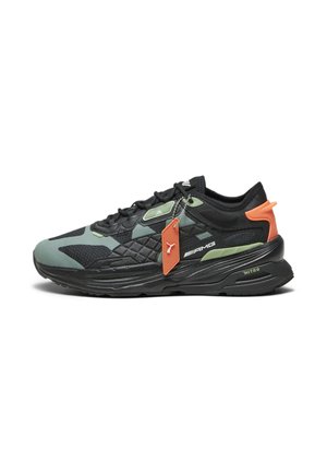 Zapatilla deportiva negra y verde con un upper texturizado, acentuada por una lengüeta en el talón de color naranja y una pequeña etiqueta naranja, con una suela gruesa negra.