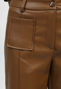 Pantalon en cuir marron avec une grande poche carrée, texture lisse et design ajusté avec une fermeture à bouton à la taille.