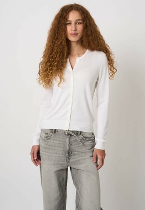 Vrouw met lang krullend haar, gekleed in een wit vest met knopen en vervaagde grijze spijkerbroek, staand tegen een effen lichte achtergrond.