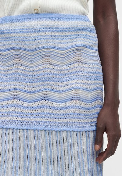 Κοντινό πλάνο ενός ατόμου που φοράει πλεκτή φούστα με μπλε και άσπρες ριγέ με κυματιστά και κάθετα σχέδια, εμφανίζοντας το δεξί του χέρι και χέρι δίπλα στη φούστα.