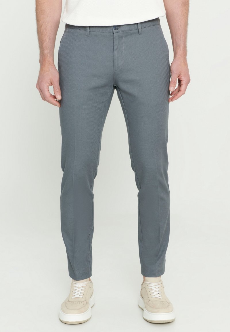 Homme portant un pantalon gris ajusté et des baskets beige, debout devant un fond clair uni.