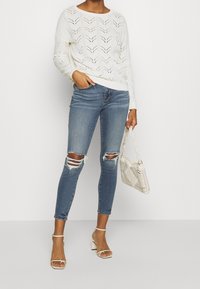 White, knit sweater with chevron pattern, paired with light blue skinny jeans featuring knee rips. Accessories include a woven bag and heels.